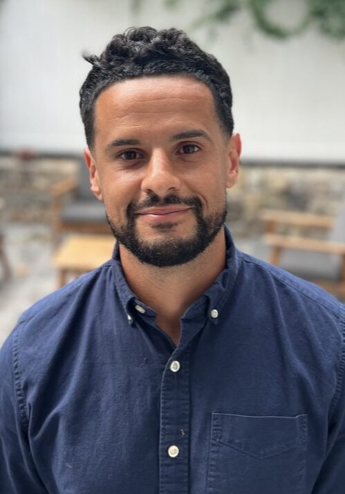 Screenshot A man with beard and blue shirt in front of a wall.